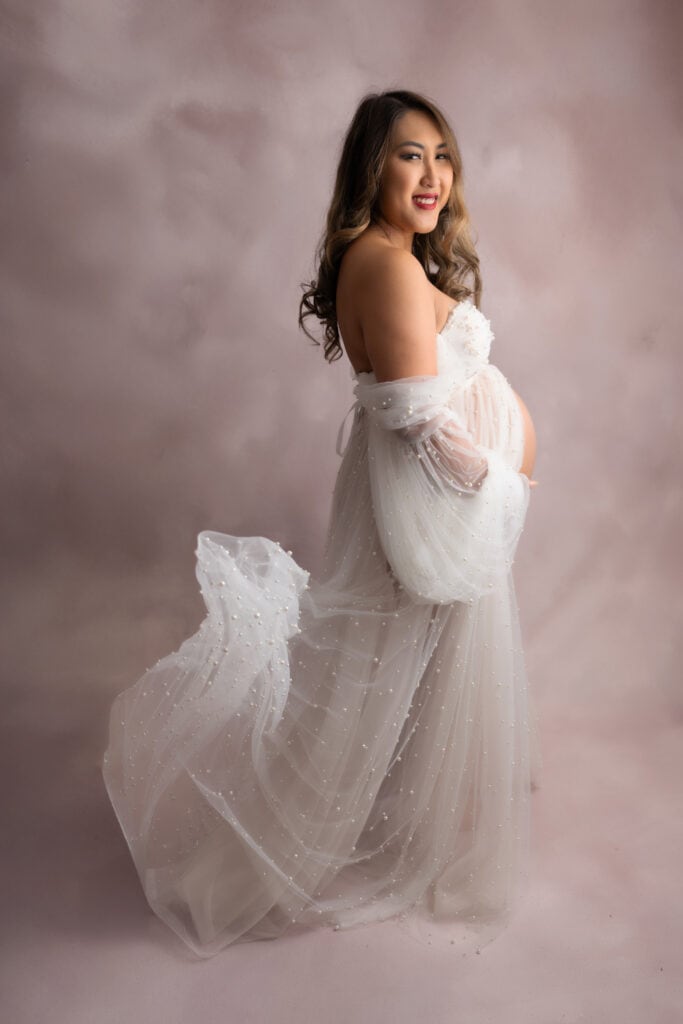 Women in a white maternity dress stands on her side as she gently touches her belly.