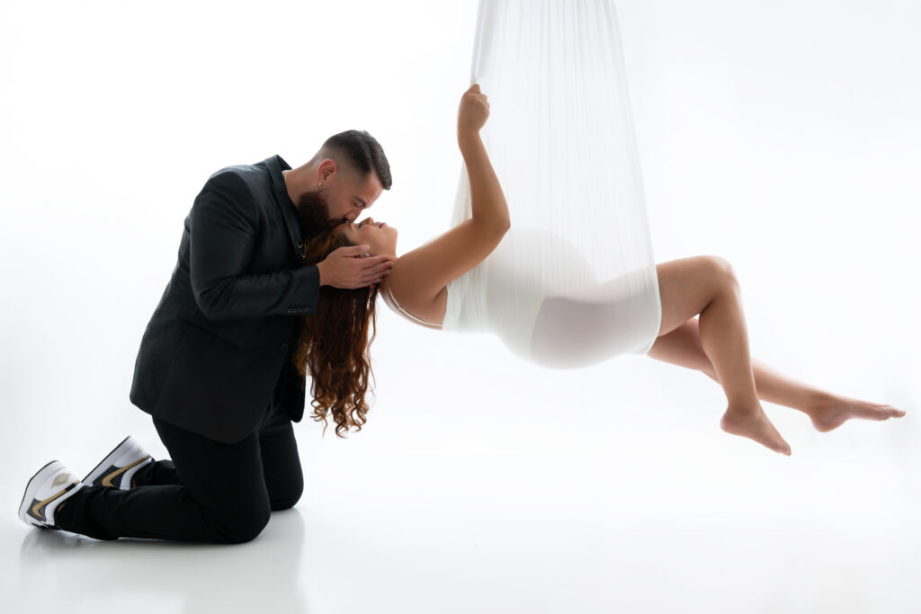 Man kneeling close to a pregnant women who is hanging from a white fabric.