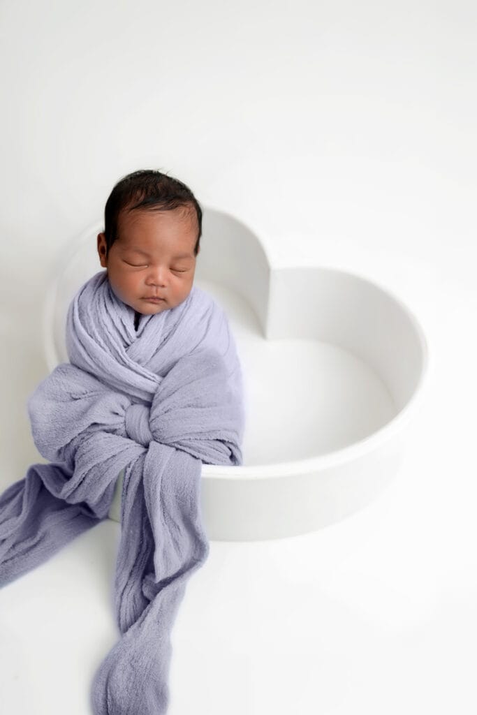 Baby in a purple wrap, tied into a bow, lays inside a heart bowl.
