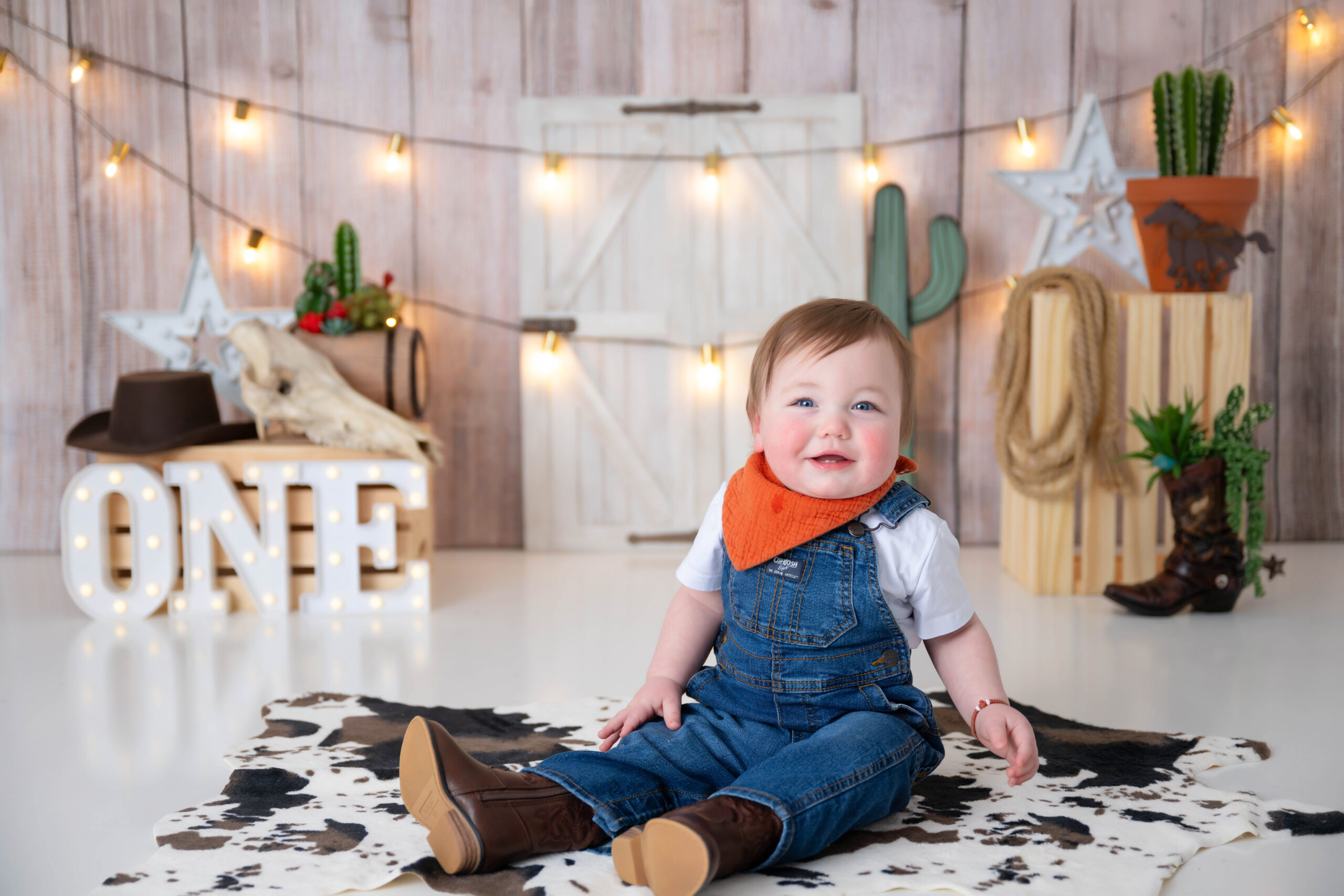 Child in overalls and boots sitting on a cow hide surrounded by western props.