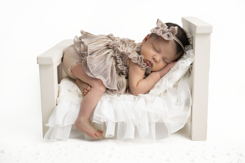 In an all white studio, baby lays on a minature bed, dressed in a neutral colored dress.
