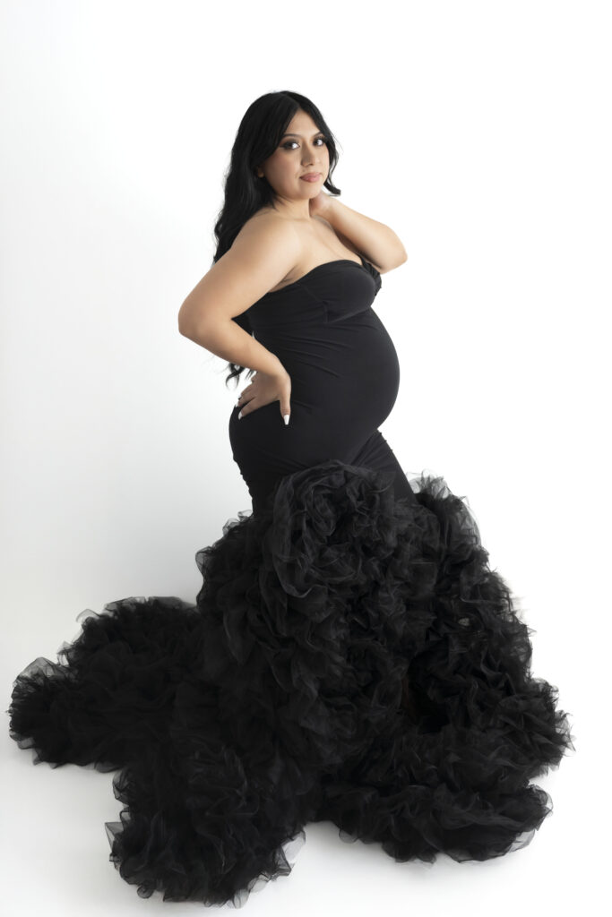 Expecting mother in a black form fitting dress stands as one hand sits on her lower back and the other caresses her neck. 