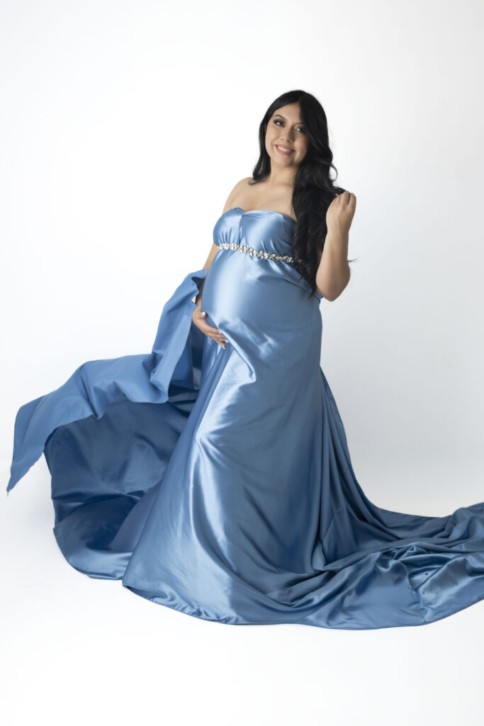 With one hand on her shoulder and another embracing her pregnant belly, a woman stands. She is draped in a silky blue fabric.