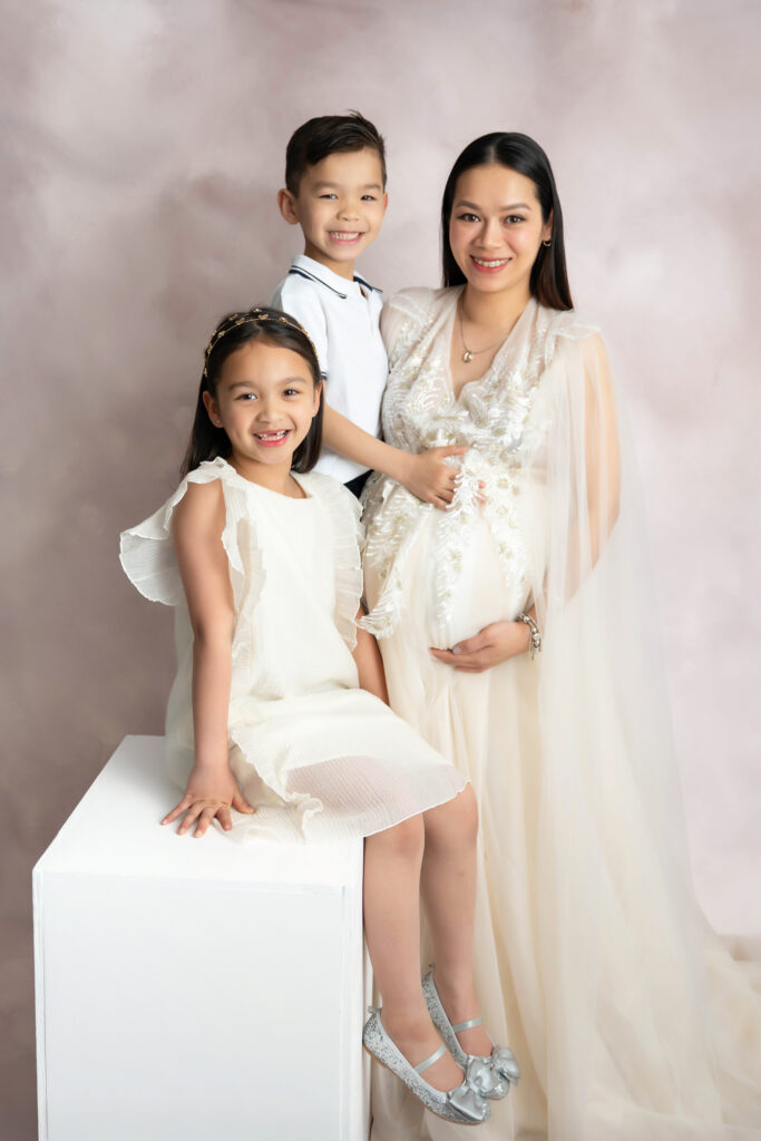 Mom holding belly surrounded by two young kids in a beige and white attire.